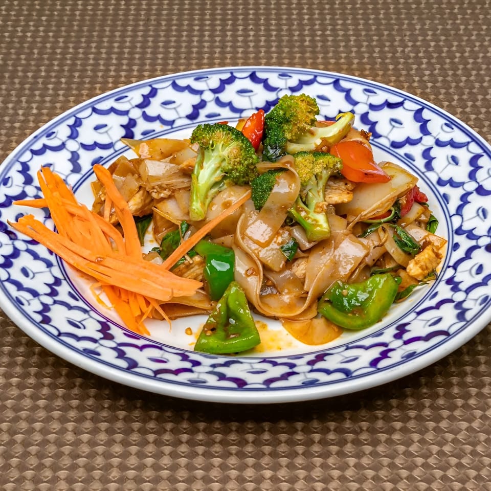 Thai Drunken Noodles with broccoli, bell peppers, carrots, and chicken on a decorative plate.