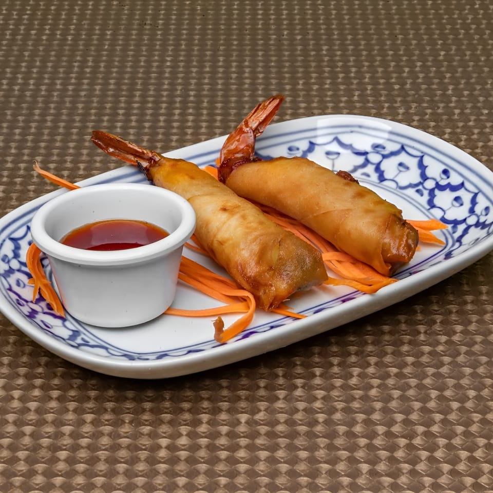 Two golden-brown shrimp spring rolls with carrot ribbons and a dipping sauce on a patterned plate.