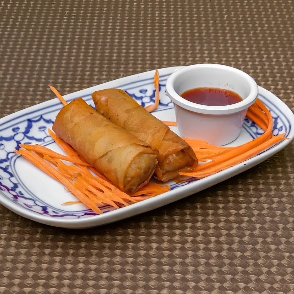 Two crispy spring rolls on a white plate with shredded carrots and a small cup of dipping sauce.