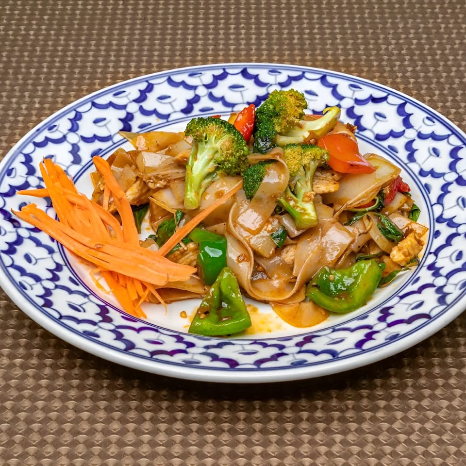 Pad See Ew with wide noodles, chicken, broccoli, bell peppers, and shredded carrots on a patterned plate.