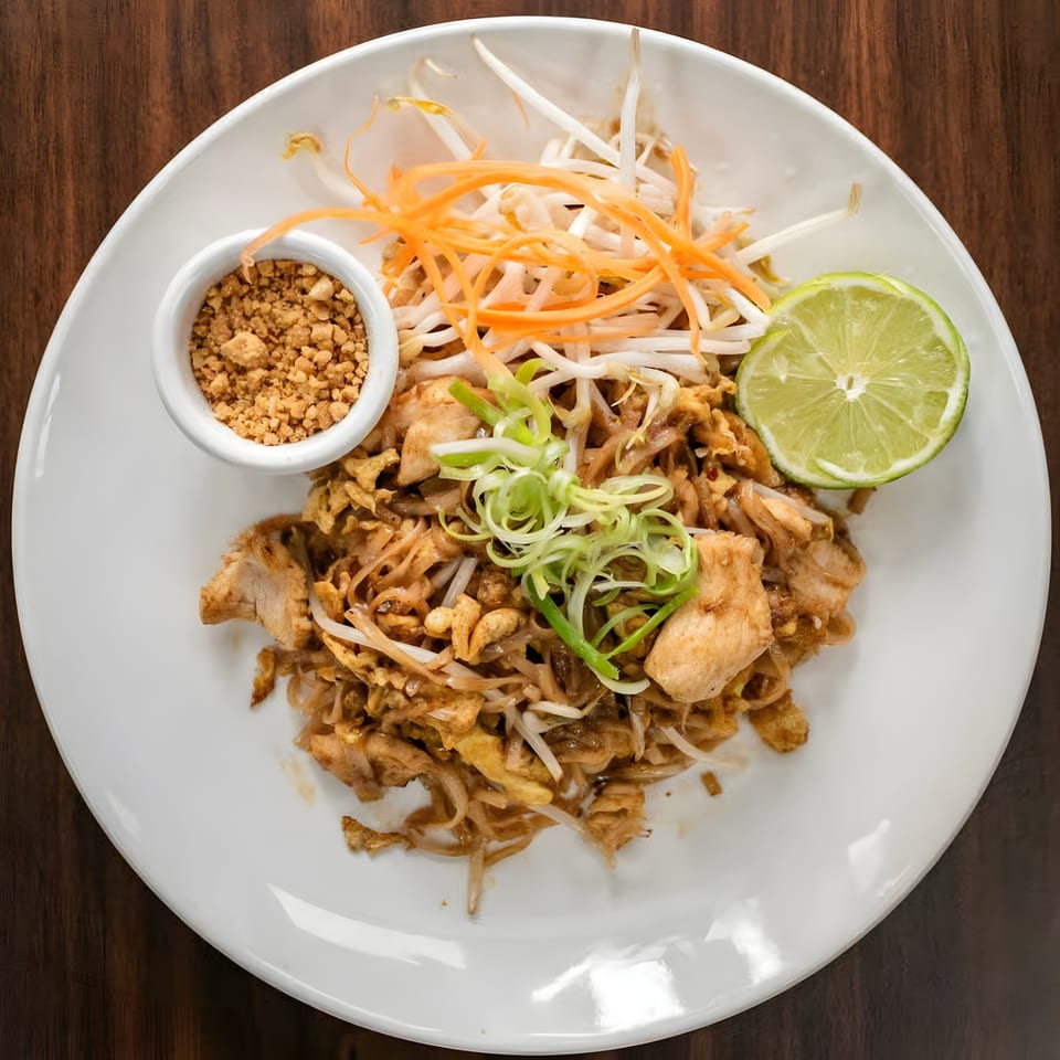 Chicken Pad Thai with bean sprouts, shredded carrots, lime, and crushed peanuts on a white plate.