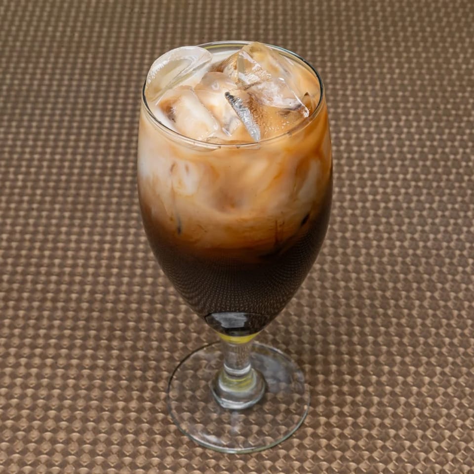 Iced coffee with cream and ice cubes in a stemmed glass on a textured brown surface.